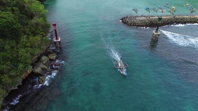 Aerial drone footage of a traditional fishing boat leaving the harbor pier and heading toward the open sea. Cinematic aerial perspective capturing the vessel departing from the dock