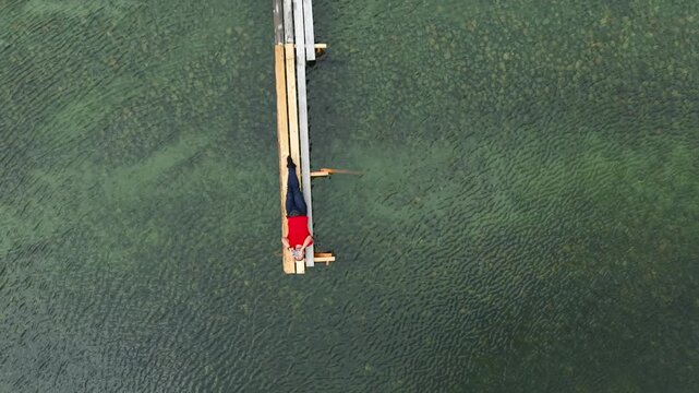 Top down drone aerial view of a male tourist in a red t-shirt lying on a wooden pier and taking a selfie as the camera ascends straight up.