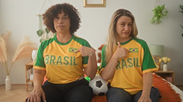 Woman friends middle age in brasil jerseys point finger while seated on bed holding a soccer ball, sharing playful expressions in a sunlit bedroom with plants and lamp; team pride.