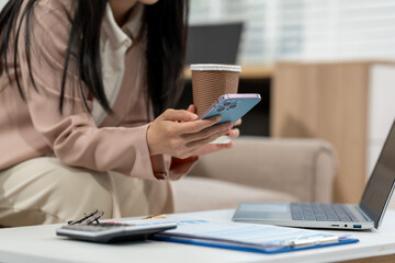 Fototapeta premium Close view of a businesswoman checking her smartphone while holding a coffee cup beside a laptop and documents in a modern office lounge.