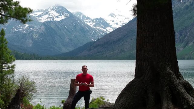 An athletic male tourist in a red t-shirt sitting on a lake shore and using a drone to take a selfie against majestic snow-capped mountains in Altai.