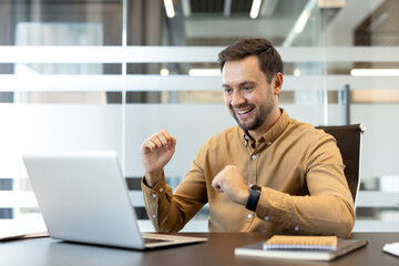 Obraz premium Young man celebrating victory looking at laptop, raising fists in a gesture of success, working remotely from a modern office environment
