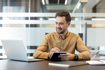 Obraz premium Young businessman engaging in productive work, taking notes in a notebook while using a laptop during a meeting or focused study session in a modern office setup