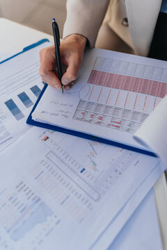 Business associates review financial reports and charts while one person writes notes with a pen. A close-up view of hands, documents and data charts during collaborative analysis.
