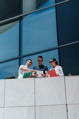 Fototapeta premium Three business associates examine paperwork together while standing on an office balcony. Colleagues collaborate and discuss documents in front of a modern glass building.