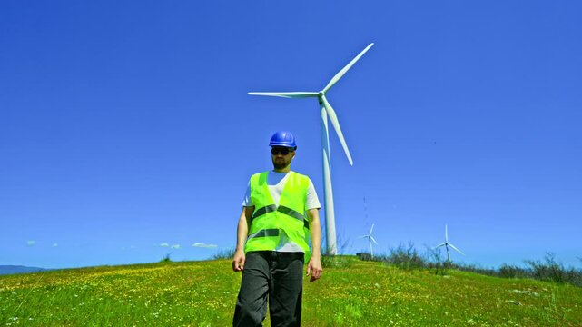 A windmill engineer stands on green grass in a rural area wearing a hard hat and safety vest. Wind turbines spin in the background on a sunny day with blue sky.