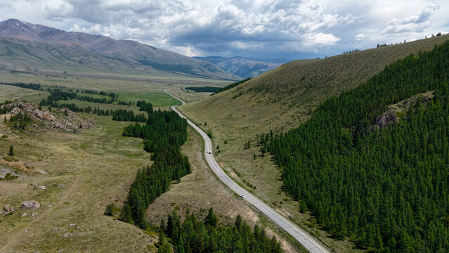 A magnificent high-angle panoramic aerial view of the scenic Chuysky Trakt highway winding through green hills towards the expansive Kurai Steppe in Altai.