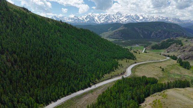 A cinematic drone revealing shot flying backwards over a winding asphalt road, uncovering an epic panorama of snowy Altai mountains.