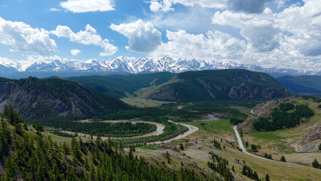 Spectacular wide aerial view of the Chuya river winding through a lush green valley with a majestic snowy mountain range in Altai.