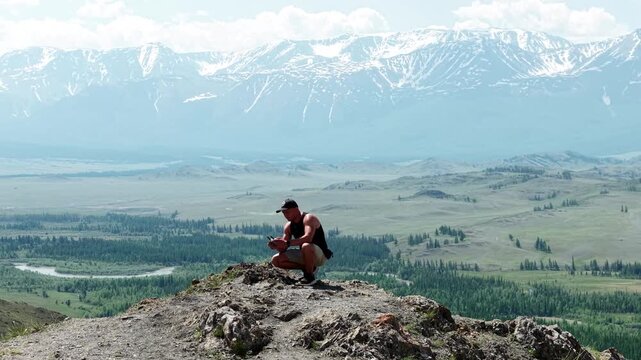 An athletic male adventurer squatting on a mountain peak and using a drone to take a panoramic selfie against a majestic snow-capped ridge.