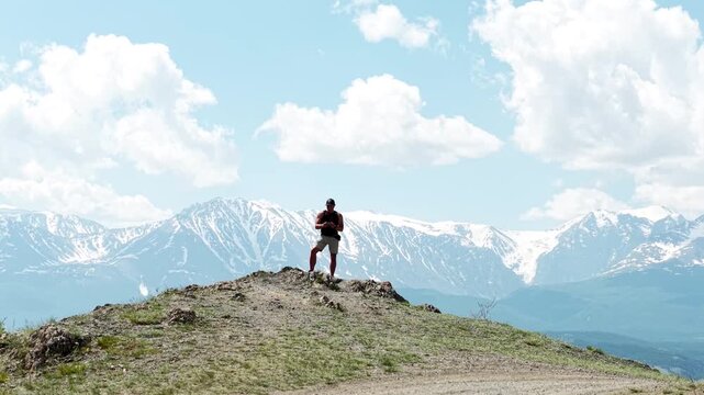 An athletic male adventurer standing on a mountain peak and using a drone to take a selfie against a majestic snow-capped ridge.