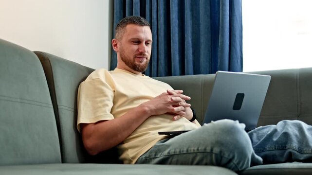 A man sits on a sofa and works on a laptop. He is engaged in tasks while surrounded by a simple living room. The afternoon light comes through a window nearby.