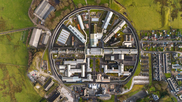 Aerial view of a circular prison complex with radiating wings surrounded by green fields and residential areas in Slough, Berkshire, United Kingdom.