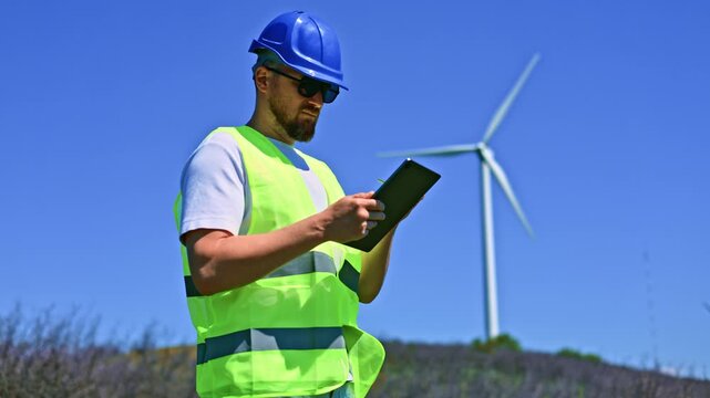 A windmill engineer stands in a bright field with a tablet in hand, observing a wind turbine as it spins. The day is sunny and the sky is clear.