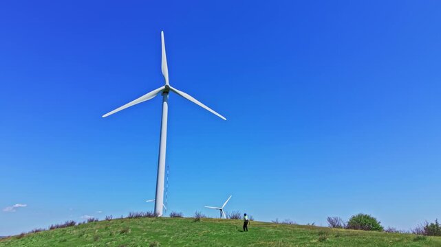 An engineer stands on a green hill in the countryside, checking the operation of windmills under a clear blue sky during the day. The scene shows multiple wind turbines in the background.