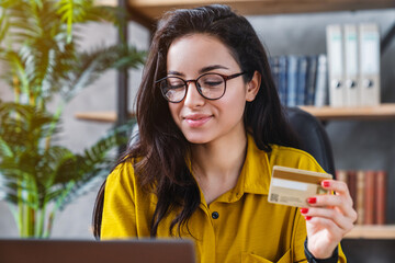 Young woman holding credit card and using laptop computer in home office, online shopping, e-commerce, internet banking and spending money concept