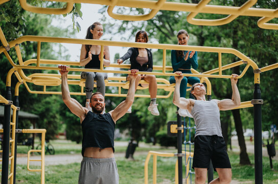 Four friends exercise on yellow monkey bars, two men performing pull-ups while women watch and clap. Group training takes place outdoors in a park, showing teamwork and fitness motivation.