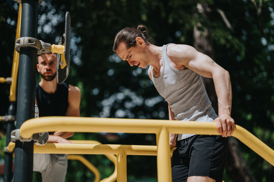 Two men work out on yellow parallel bars at an outdoor gym, one performing dips while the other watches.