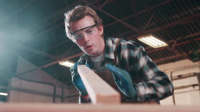 Carpenter, cleaning and blow dust with plank in workshop, furniture manufacturing or production process. Woodwork, man and remove sawdust, craftsmanship and wipe shavings in carpentry industry