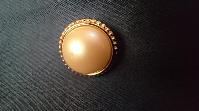 Elegant macro close-up of a luxurious cream pearl button with a decorative gold frame, set against a dark textured fabric background, ideal for fashion design, formal wear, and high-end clothing detai