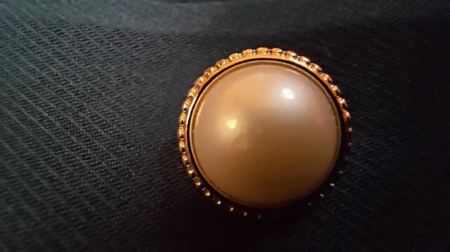 Elegant macro close-up of a luxurious cream pearl button with a decorative gold frame, set against a dark textured fabric background, ideal for fashion design, formal wear, and high-end clothing detai