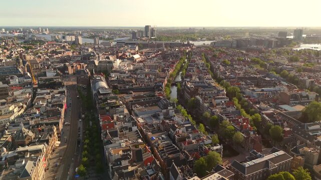 Drone follows a tree lined canal above Amsterdam flying forward toward the IJ harbor and city skyline in warm light
