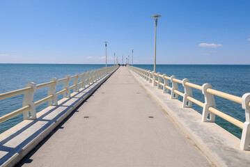 Obraz premium Long concrete pier extending into a calm blue sea beneath a clear sky, featuring strong leading lines, symmetry, and a peaceful coastal atmosphere with ample copy space.