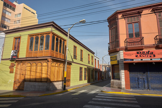 A charming historic corner in Tacna, Peru, showcases classic colonial architecture with a striking blue upper facade, wooden balcony, and weathered stone masonry below. Traditional wrought iron railin