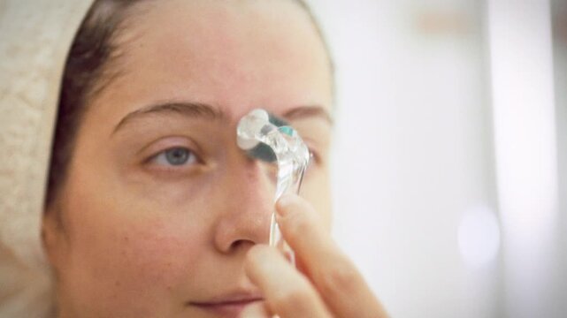 In a bathroom, a girl in a white robe with a towel on her head uses a derma roller for microneedling before a mirror, a skin-care step that stimulates renewal and boosts collagen manufacture