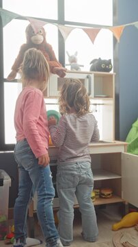 Girl and younger child play at a wooden toy kitchen, holding plates and a stuffed monkey beside a window with bunting in a building; joy.