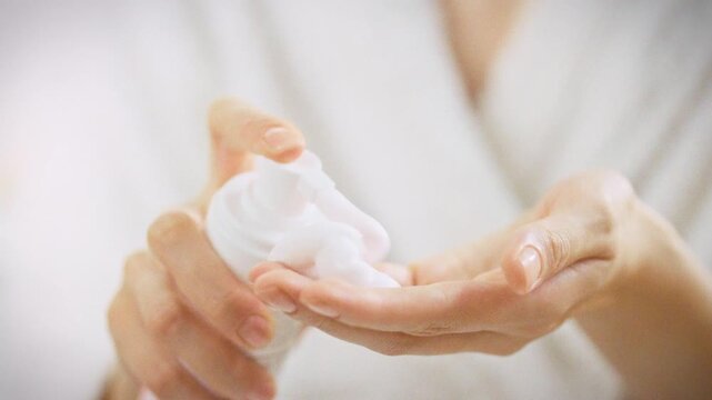 In a bright bathroom vanity, a girl gently squeezes white foam from a skincare bottle onto her hands, preparing to cleanse her face with a soothing facial care routine, emphasizing daily skin health