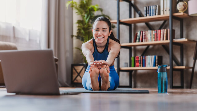 Sporty young woman stretching on mat while watching online tutorial on laptop at home, fit female exercising indoors, home workout, fitness and healthy lifestyle concept