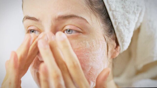 In bright bathroom, a young woman with a towel wrapped around her head spreads white cleansing foam across her face, a daily ritual that highlights her focus on cosmetic care and a healthy lifestyle