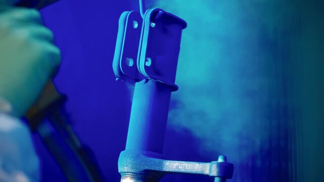 Powder coating gun spraying blue powder onto suspended metal automotive bracket inside industrial spray booth with bright particles, precise finish, and controlled surface coverage