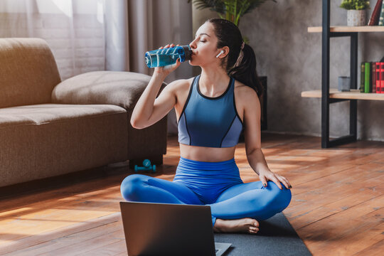 Fit woman in sportswear sitting on mat and drinking water from bottle while using laptop in living room at home, fitness, wellness and healthy lifestyle concept