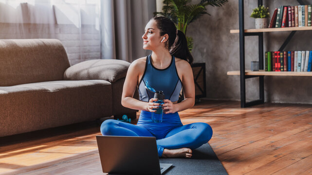 Dreaming sporty woman in earphones sitting on mat with bottle of water while using laptop in living room at home, fitness, wellness and healthy lifestyle concept
