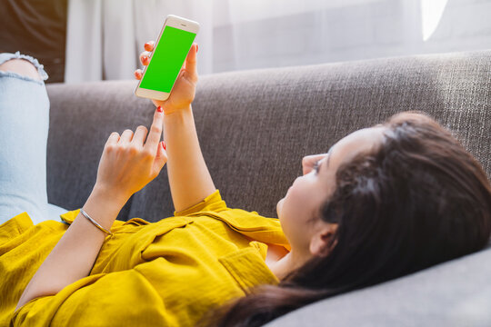 Young woman using smartphone with mockup screen while sitting on sofa in living room at home, female browsing mobile app, social media and cozy lifestyle concept