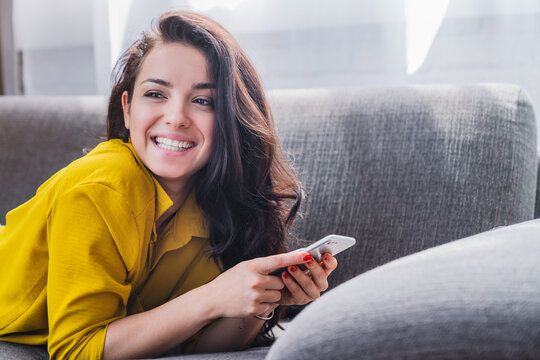 Young woman smiling and using mobile phone while lying on sofa in living room, happy female texting online, social media and cozy home lifestyle concept