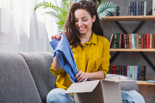Young happy woman sitting on sofa unpacking parcel delivery box with clothes at home, satisfied customer enjoying online shopping, e-commerce and home delivery concept