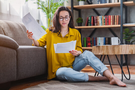 Young woman looking at bills while sitting on floor in living room, managing home budget, household expenses, financial planning and money saving concept