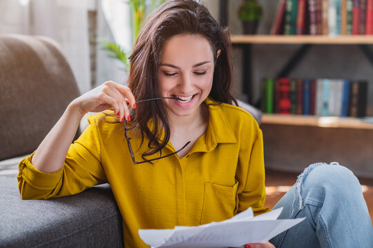 Young smiling woman sitting on floor and checking bills in living room, happy female managing household budget, financial planning and home expenses concept
