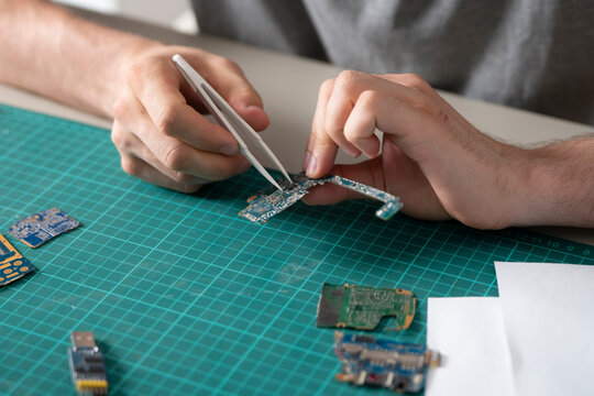 Hands carefully handle a circuit board using tweezers in an electronics workshop. Tools and components are nearby