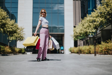 Fototapeta premium A young woman stands outside Diamond Mall holding several colorful shopping bags. She poses confidently after a day of shopping in a modern outdoor mall.