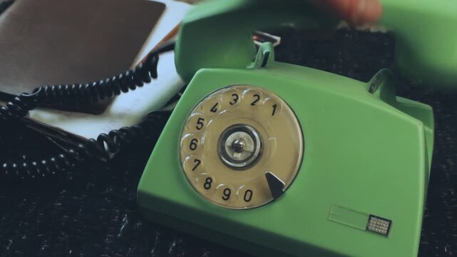 Close up of a hand using a pen to turn the dial on a vintage green rotary telephone, demonstrating an outdated method of communication and retro office telephony with a notebook in the background