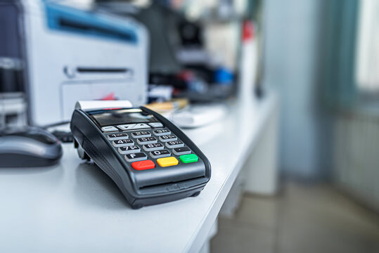 A black credit card terminal sits on a white office desk next to a computer mouse and a printer.