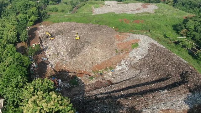 Aerial drone footage of two excavators operating on the active landfill cell at a waste disposal site. 
The heavy machinery performs waste compaction and land preparation in a municipal solid waste