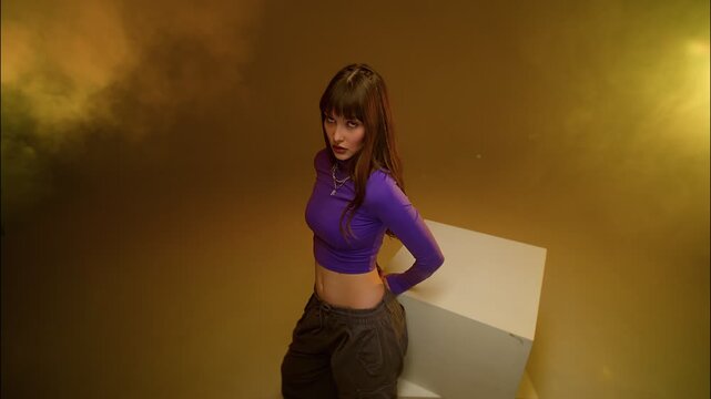 Young woman leaning beside white prop in warm gold studio with purple top and serious editorial mood