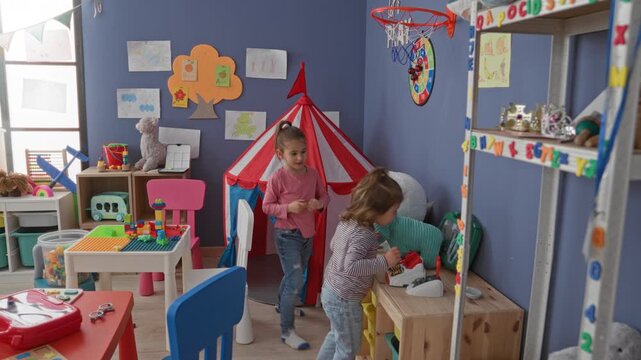 Girl holds stuffed toy and reaches for a toy phone by a red play tent in a children's playroom inside a building; playful friendship.