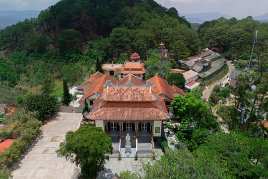 Traditional Southeast Asian temple buildings with terra cotta roofs and a white statue stand on a green mountain slope under bright daytime skies