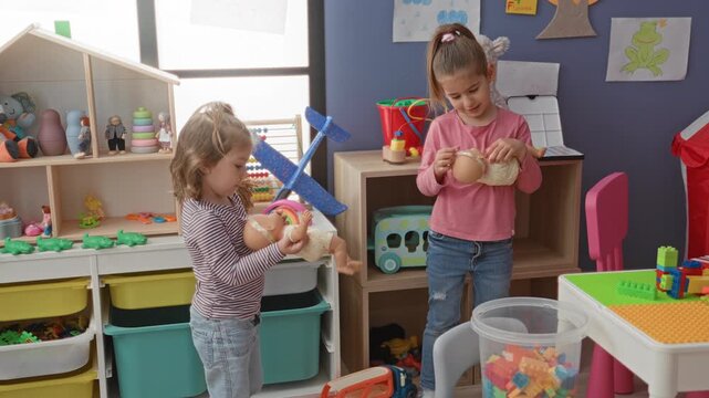 Two girls cradle baby dolls facing each other in a preschool playroom with toy shelves, blocks and storage bins; playful care.